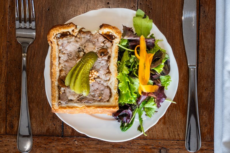 Une tranche de pâté en croûte maison, cornichons et salade, sur une assiette blanche.